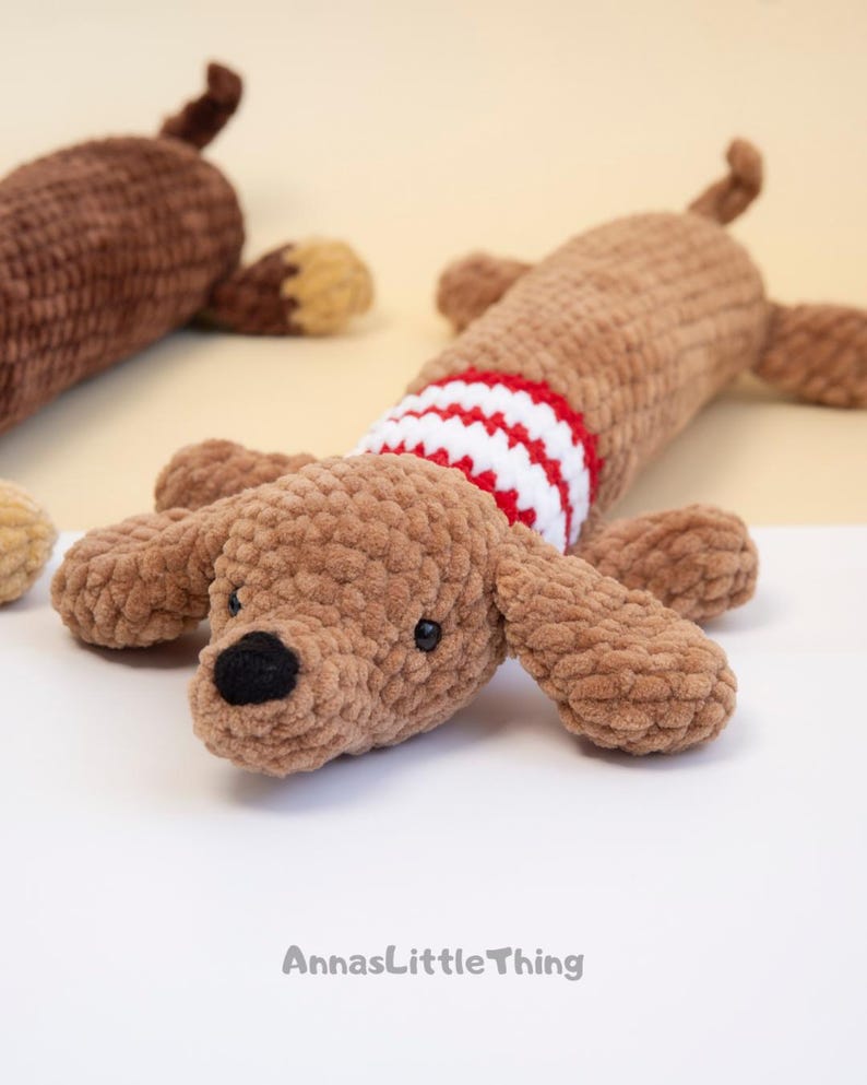 May include: Two crocheted plush dachshund toys made from soft brown yarn. The foreground dog features a red and white striped sweater pattern around its torso, black yarn eyes, and a black nose. The text AnnasLittleThing is printed at the bottom.