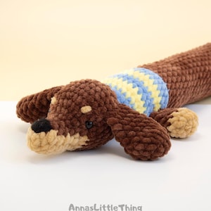 May include: A brown crocheted dachshund toy with a tan muzzle, black nose, and small black eyes. The dog wears a blue and yellow striped sweater around its midsection. The text AnnasLittleThing is printed in gray at the bottom of the image.