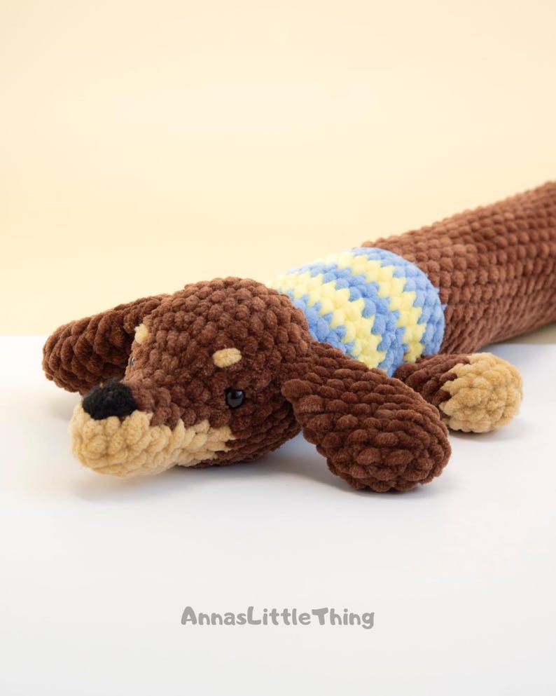 May include: A brown crocheted dachshund toy with a tan muzzle, black nose, and small black eyes. The dog wears a blue and yellow striped sweater around its midsection. The text AnnasLittleThing is printed in gray at the bottom of the image.