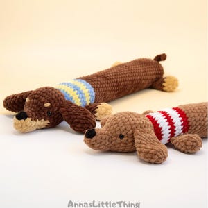 May include: Two crocheted dachshund toys made of soft brown yarn. The back dog wears a blue and yellow striped sweater, while the front dog wears a red and white striped sweater. Both have black noses and small dark eyes. Text reads AnnasLittleThing.