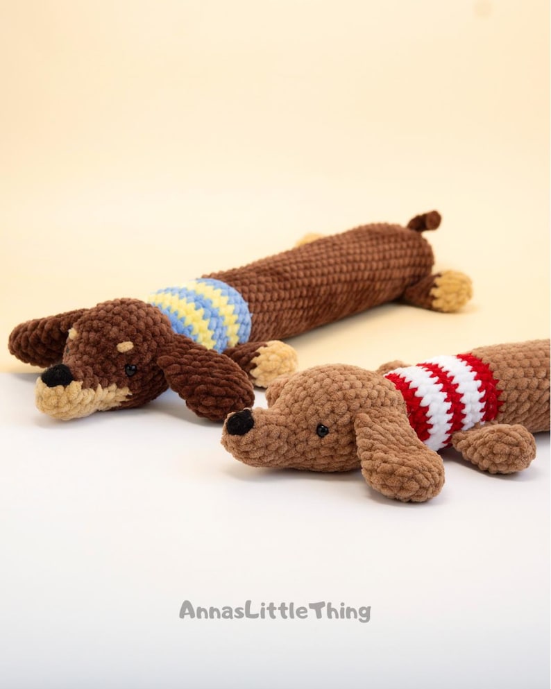 May include: Two crocheted dachshund toys made of soft brown yarn. The back dog wears a blue and yellow striped sweater, while the front dog wears a red and white striped sweater. Both have black noses and small dark eyes. Text reads AnnasLittleThing.