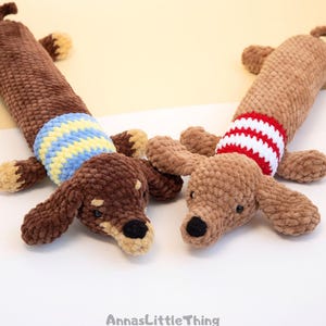 May include: Two plush dachshund toys made of soft brown yarn. The left dog has a blue and yellow striped sweater, while the right dog wears a red and white striped sweater. Both have black noses and small ears. The text AnnasLittleThing is visible.