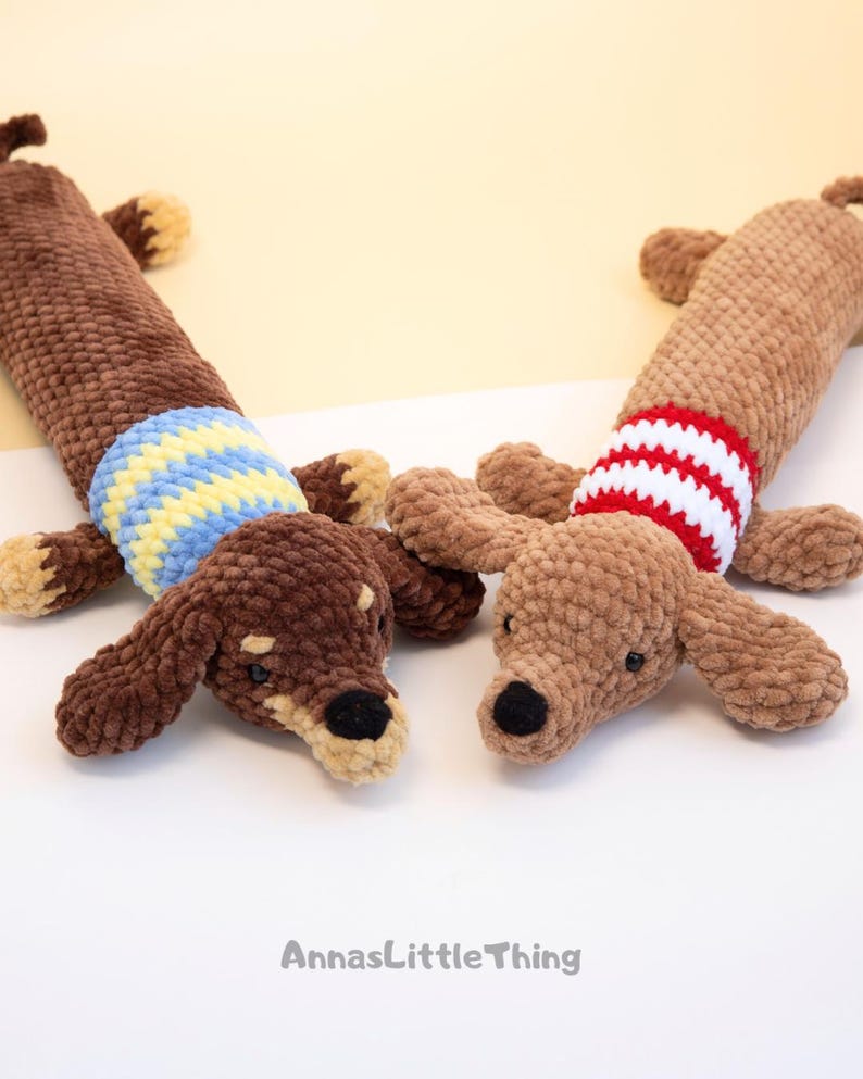 May include: Two plush dachshund toys made of soft brown yarn. The left dog has a blue and yellow striped sweater, while the right dog wears a red and white striped sweater. Both have black noses and small ears. The text AnnasLittleThing is visible.