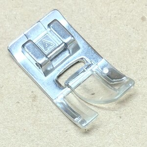 May include: A silver sewing machine presser foot with a clear plastic section. The foot has an "A" marking and is designed for sewing. The item is shown on a tan surface.