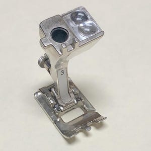 May include: A silver sewing machine presser foot with a number 3 on the side. The foot has a rectangular base with a curved front edge and a small opening. The top has two clear plastic pieces.