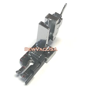 May include: A silver-colored metal sewing machine presser foot with a hinged design and a vertical shank attachment. The component features a central needle opening and the text SEWVACUSA printed in red letters across the side.