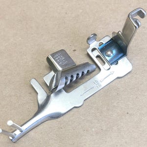 May include: Silver sewing machine foot with a ruler marked 15/16 inches. The foot has a small metal plate with the text "0.813 5/16 0.701 1/2 0.550".