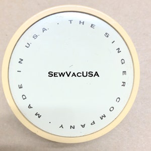 May include: A white round lid with black text that reads "Made in U.S.A. The Singer Company SewVacUSA".