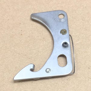 May include: A silver-colored metal part with a curved design, featuring a hook-like end and several holes. A small spring is attached to the side. The object appears to be a mechanical component.