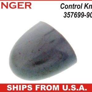 May include: A gray Singer Control Knob, model 357699-905, with a rounded, teardrop shape. The knob has a smooth surface with subtle color variations. The Singer logo is visible in the upper left corner.