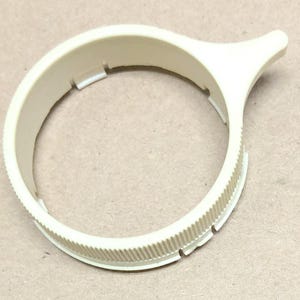 May include: A cream-colored, circular plastic component with a textured outer edge and a small, protruding handle. The ring has several rectangular cutouts. The object is likely a dial or control knob for a machine or device.