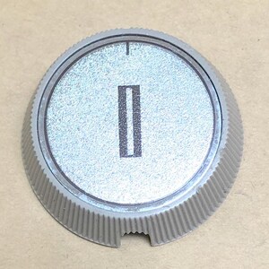 May include: A circular light gray control knob with a textured, ribbed outer edge and a flat, metallic silver center face. A single vertical black rectangular indicator mark is printed in the middle, with a small black tick mark at the top edge.