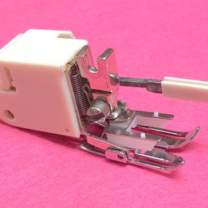 May include: A white plastic sewing machine foot with a metal foot plate and a white plastic guide. The foot is designed for sewing on thick fabrics.