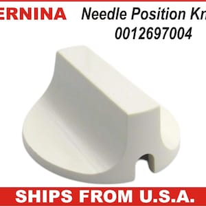 May include: A white Bernina Needle Position Knob with the number 0012697004 printed on it. The knob has a curved base and a rectangular top with a notch on one side. The Bernina logo is in red.