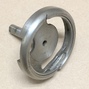 May include: A vintage, silver-toned sewing machine part, likely a bobbin case, with a circular design and a protruding shaft. The metal component shows signs of wear, suggesting its age and use. The part has a curved opening and a central hole.