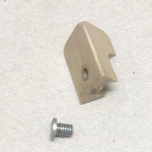 May include: A tan-colored plastic component with a curved shape and a hole, next to a silver metal screw. The plastic piece has a slightly rough texture. The screw has a threaded shaft and a flat head.