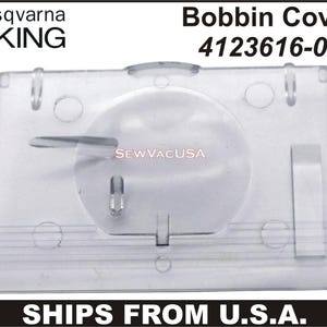 May include: Clear plastic Husqvarna Viking bobbin cover. The cover has a rectangular shape with rounded edges and a central circular area. The text "Bobbin Cover 4123616-01" is printed at the top. The logo "SEW VAC USA" is also visible.