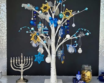 May include: A white decorative birch tree with blue and gold ornaments, including Star of David shapes and blue bows. A silver-toned menorah stands to the left, and a glass jar filled with gold-colored chocolate coins sits to the right.