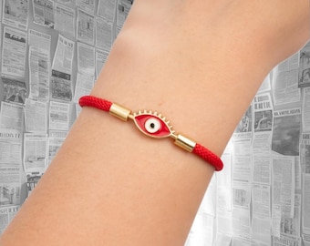 May include: A red cord bracelet featuring a central gold-tone metal charm shaped like an eye. The charm has a red enamel iris, a white sclera, and a black pupil. The cord is secured on both sides of the eye charm by small gold-tone metal cylinders.