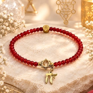 May include: A beaded bracelet featuring small, faceted, translucent red beads. The bracelet includes a gold-toned metal charm shaped like a Star of David with a central clear stone, a dangling gold-toned Hebrew Chai symbol, and a small round gold bead.
