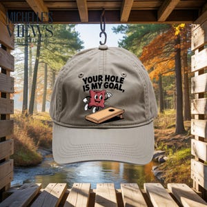 May include: A tan baseball cap with the text "YOUR HOLE IS MY GOAL," featuring a cartoon cornhole bag and board. The cap is hanging from a hook, with a scenic background of a river and trees.