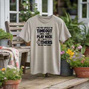 May include: A light tan t-shirt with the text "I'VE PUT MYSELF IN TIMEOUT UNTIL I CAN PLAY NICE WITH OTHERS THIS MAY TAKE AWHILE" in black. The shirt is displayed outdoors.