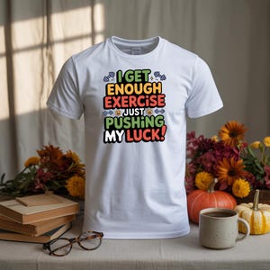 May include: White t-shirt with the text "I GET ENOUGH EXERCISE JUST PUSHING MY LUCK!" in a colorful, playful font. The design includes dumbbell and emoji graphics. The shirt is displayed on a table with fall-themed decorations.