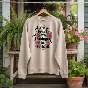 Puede incluir: Sudadera beige con las palabras "VERY FERAL VERY UNHINGED VERY BATSHIT" en fuente gótica negra, rodeada de rosas rosas y murciélagos. La sudadera está colgada de una percha de madera.