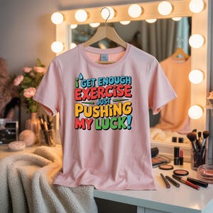 May include: A light pink t-shirt with the text "I get enough exercise just pushing my luck!" in colorful, bold letters. The shirt is hanging on a wooden hanger in front of a lighted vanity mirror.