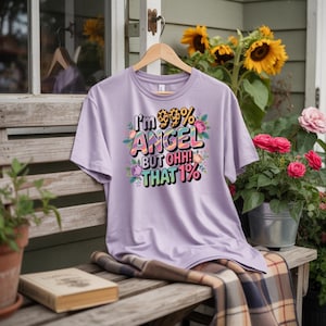 Könnte beinhalten: Ein helllila T-Shirt mit dem Text "I'm 99% ANGEL BUT OHH! THAT 1%" in einem farbenfrohen, floralen Design. Das Shirt hängt an einem Holzbügel vor dem Hintergrund von Sonnenblumen und Rosen.