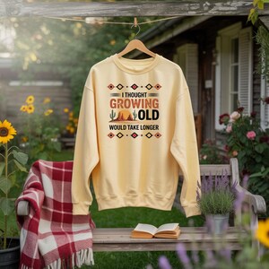Puede incluir: Una sudadera amarilla clara con el texto "I thought growing old would take longer" en un diseño de estilo suroeste. El diseño incluye un cactus, un sol y formas geométricas en marrón, naranja y rojo.