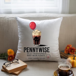 May include: White square pillow featuring a graphic of a root beer float with a red balloon. The text reads "Pennywise Old Fashioned Root Beer Floats Derry's Darkest Delight" with a smiling clown face.