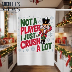 May include: White kitchen towel with the text "NOT A PLAYER I JUST CRUSH A LOT" in green and red, with snowflake accents and a cartoon nutcracker illustration. The towel has a decorative border.