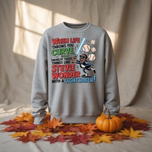 Puede incluir: Sudadera gris con el texto "WHEN LIFE THROWS YOU CURVE BALLS SWING AT THOSE THINGS LIKE STEVIE WONDER WITH A LIGHTSABER!". El diseño muestra a Stevie Wonder con un sable de luz golpeando pelotas de béisbol.