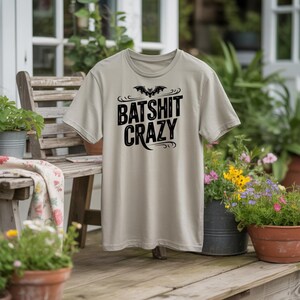 Puede incluir: Camiseta de color beige claro con la frase "BATSHIT CRAZY" en negro y un diseño de murciélago. La camiseta se muestra al aire libre, con un banco de madera y plantas en macetas en el fondo.