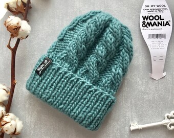 May include: A mineral blue knit beanie featuring a thick ribbed cuff and a vertical cable knit pattern. A black rectangular tag with white text reading Klybo_ok is attached to the side. The beanie is made from 100% Peruvian wool.