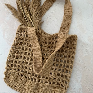 May include: A brown crocheted tote bag with a single strap. The bag is made of a thick, textured yarn and has a large, open weave. The bag is empty and is sitting on a white surface.