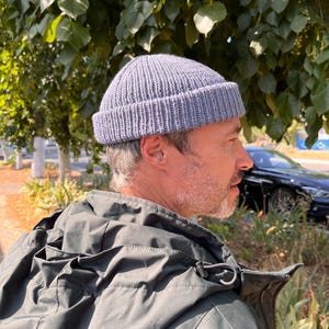 Può includere: Un berretto grigio a coste. Il cappello è realizzato in un materiale morbido e lavorato a maglia. Il cappello è indossato da una persona. La persona indossa una giacca verde scuro. Lo sfondo include fogliame verde e un'auto nera.