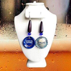 May include: A pair of earrings featuring a dark brown bead, a blue bead, and a bottle cap with the text "Samuel Adams". The other earring features a dark brown bead, a blue bead, and a bottle cap with the text "Cheers!"