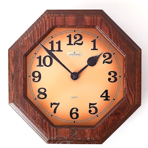Pode incluir: Um relógio de parede octogonal vintage com uma moldura de madeira escura. O mostrador do relógio é de cor laranja clara com algarismos e ponteiros pretos. O relógio diz "Junghans" e "quartz".