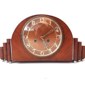 May include: A vintage wooden mantel clock with a round, glass-faced clock face with Roman numerals. The clock has a dark wood finish and a decorative design.