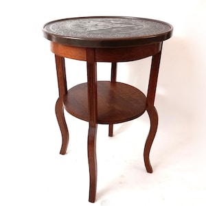 Op de afbeelding: Een ronde houten bijzettafel met een decoratief metalen blad en een lager plankje. De tafel heeft drie gebogen poten en is afgewerkt met een donkere houtkleur.