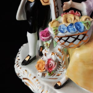 May include: A porcelain figurine depicting a couple with a basket of flowers. The man wears black pants, a white coat with gold trim, and black shoes. The woman holds a basket of colorful roses. The base is white with gold accents.