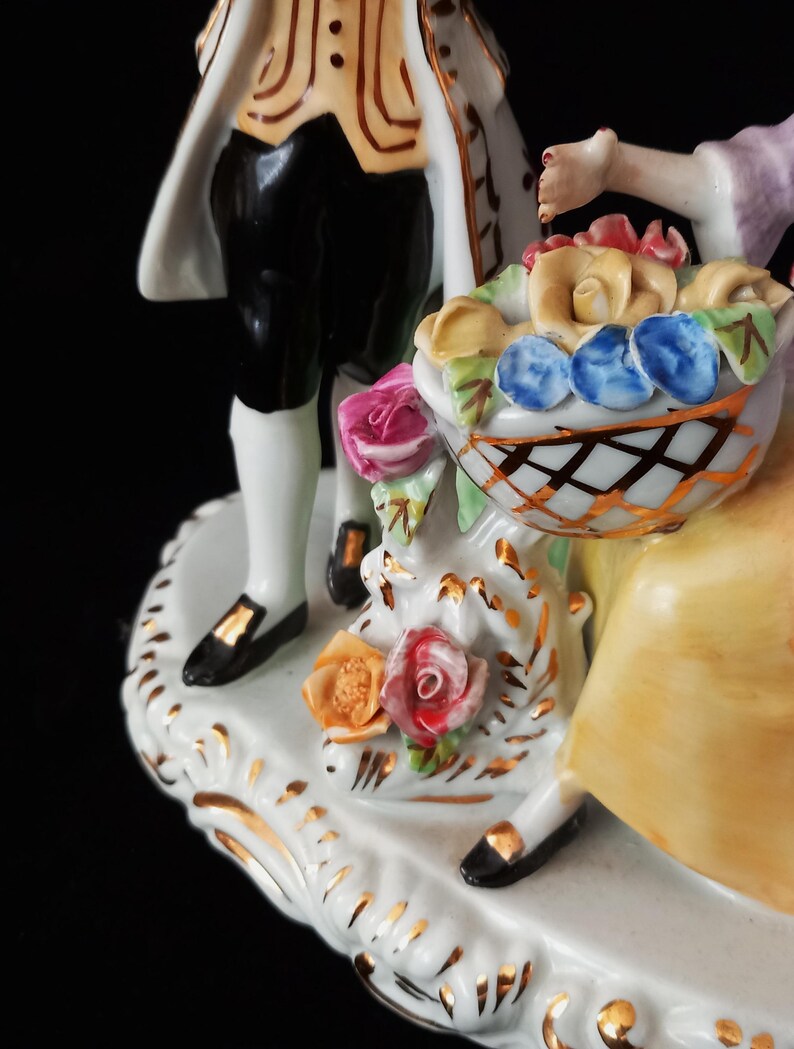May include: A porcelain figurine depicting a couple with a basket of flowers. The man wears black pants, a white coat with gold trim, and black shoes. The woman holds a basket of colorful roses. The base is white with gold accents.