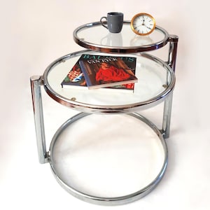 May include: A tiered side table with a polished chrome frame and two circular glass shelves. The top shelf holds a gray ceramic mug and a small gold-rimmed analog clock. The lower shelf displays two books titled Balthus and Van Eyck.