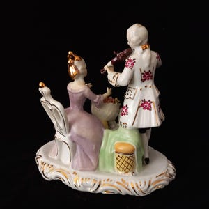 May include: A porcelain figurine depicting a man playing a violin for a woman seated in a chair. The man wears a white coat with red floral accents. The woman wears a lavender dress. The base is white with gold trim.