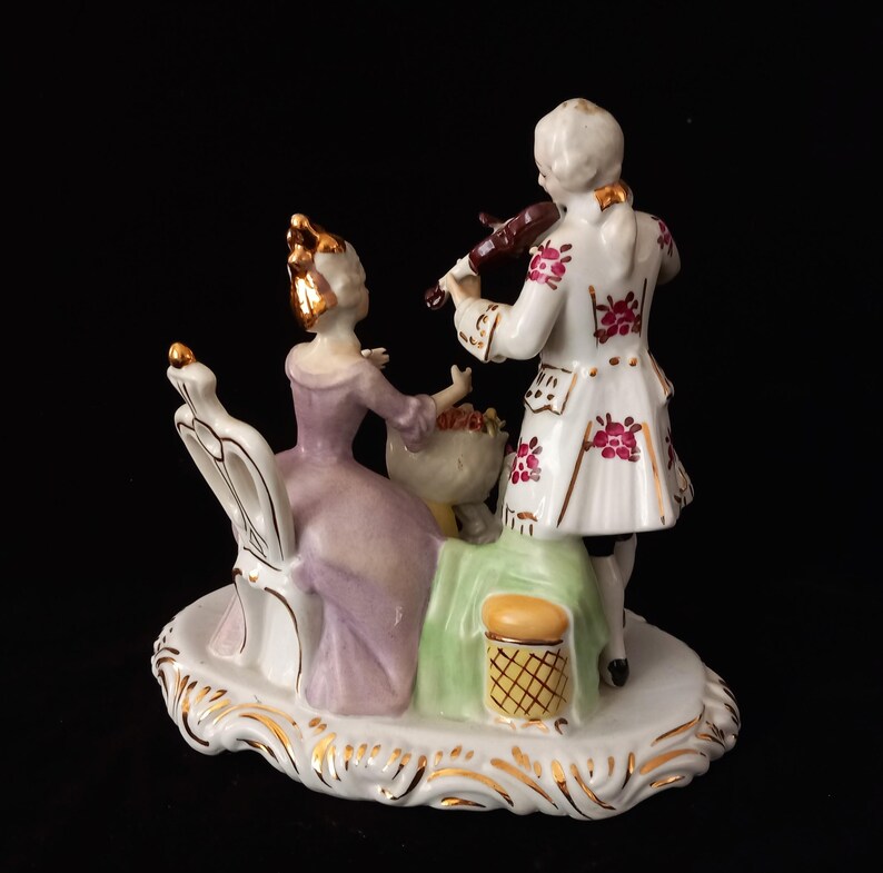 May include: A porcelain figurine depicting a man playing a violin for a woman seated in a chair. The man wears a white coat with red floral accents. The woman wears a lavender dress. The base is white with gold trim.