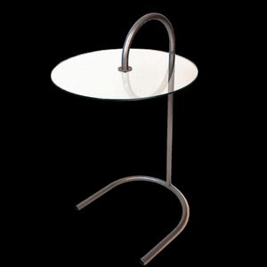 May include: A silver metal side table with a round glass top and a curved handle. The table has a simple, modern design.