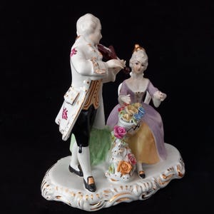 May include: A porcelain figurine depicting a man playing a violin and a woman seated with a basket of flowers. The man wears a white coat with gold trim and black pants. The woman is in a purple dress. The base is white with gold accents.