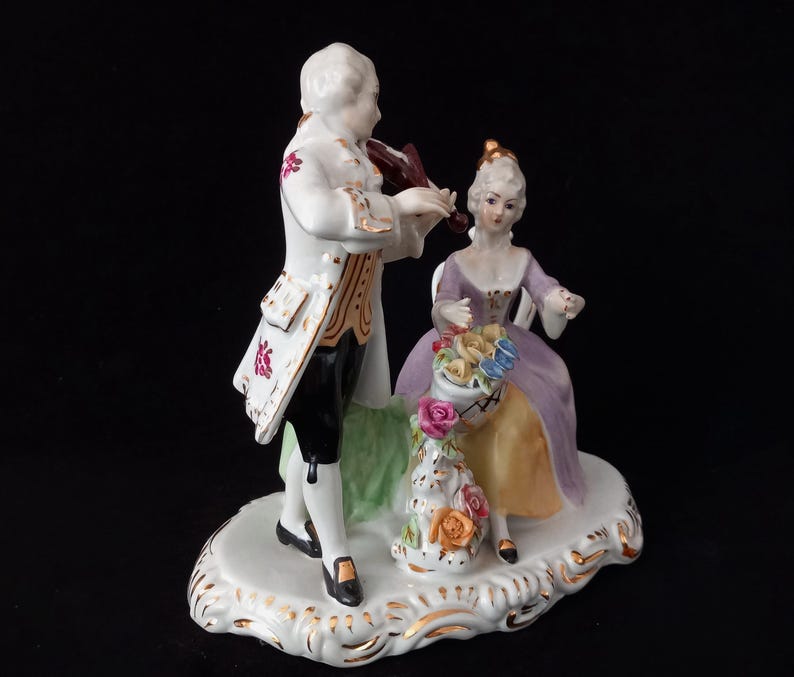 May include: A porcelain figurine depicting a man playing a violin and a woman seated with a basket of flowers. The man wears a white coat with gold trim and black pants. The woman is in a purple dress. The base is white with gold accents.